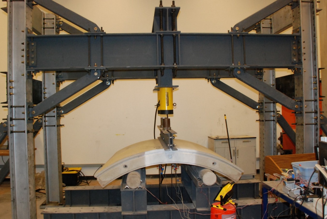 A curved precast concrete tunnel segment resting on two roller supports inside a steel load frame, with a hydraulic ram pressing on its crown