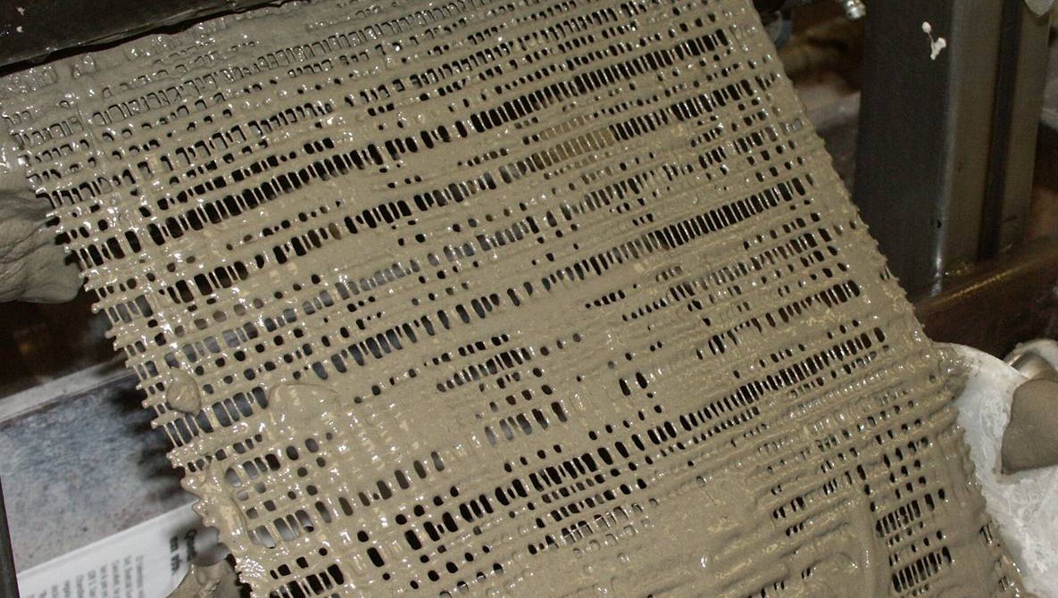 Open-mesh fabric coated with wet grey cement slurry, hanging from the rollers of a laboratory machine